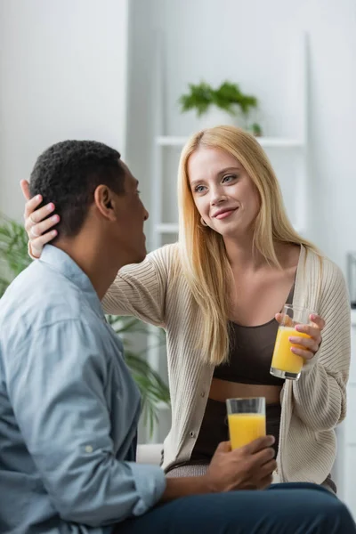 Compiaciuta giovane donna che tiene in mano un bicchiere di succo d'arancia fresco e tocca la testa dell'uomo afroamericano in cucina — Foto stock