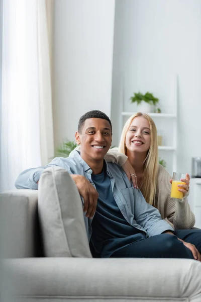 Mujer rubia con un vaso de jugo de naranja fresco sentada en el sofá cerca de novio afroamericano y sonriendo a la cámara - foto de stock