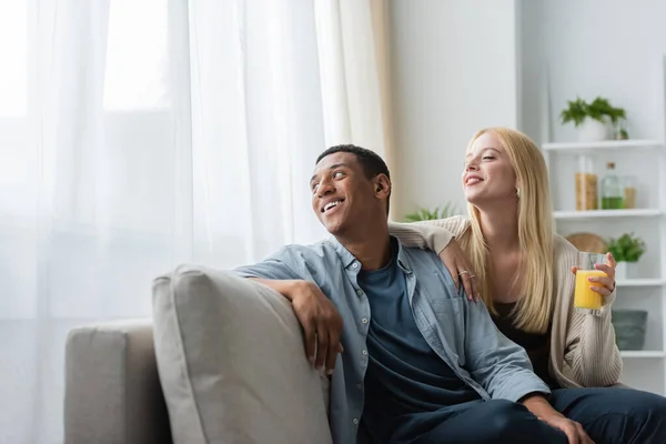 Blonde woman with orange juice and smiling african american man sitting on sofa in kitchen and looking away — Stock Photo