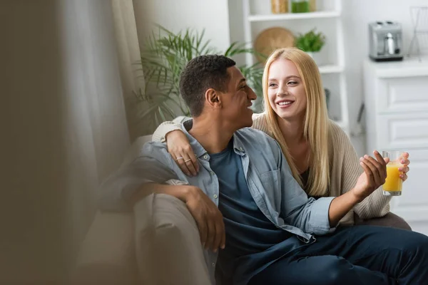 African american man and blonde woman with orange juice talking on couch in kitchen — Stock Photo