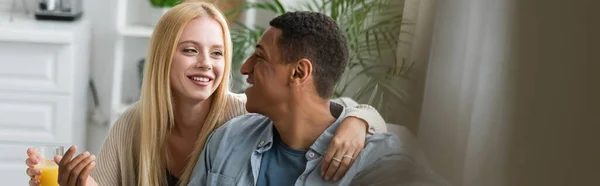 Smiling blonde woman holding orange juice and hugging african american boyfriend in kitchen, banner — Stock Photo