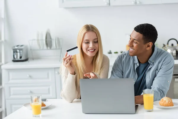 Happy young woman holding credit card and pointing at laptop near smiling african american boyfriend and breakfast in kitchen — Stock Photo