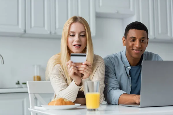 Blonde woman holding credit card near african american boyfriend and laptop in kitchen — Stock Photo