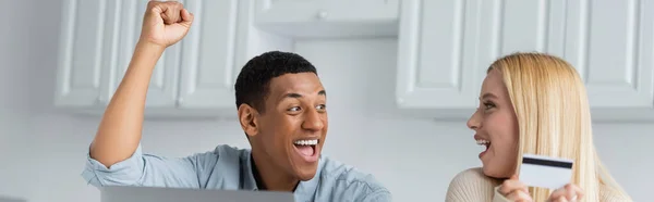 Homem americano africano alegre mostrando gesto de sucesso perto da namorada loira com cartão de crédito, banner — Fotografia de Stock