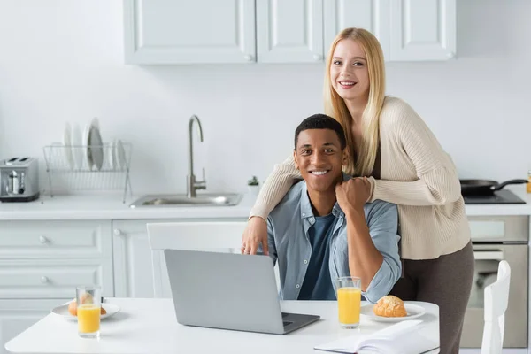 Веселая многонациональная пара, держащаяся за руки и улыбающаяся перед камерой возле ноутбука и завтрака на кухонном столе — стоковое фото