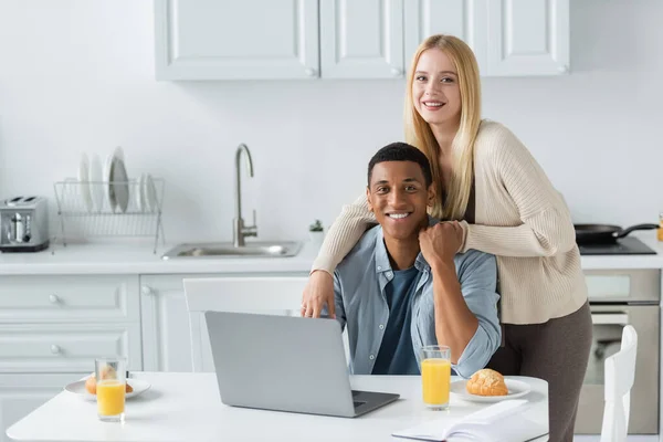 Joyeux couple multiethnique tenant la main et souriant à la caméra près de l'ordinateur portable et le petit déjeuner sur la table de cuisine — Photo de stock