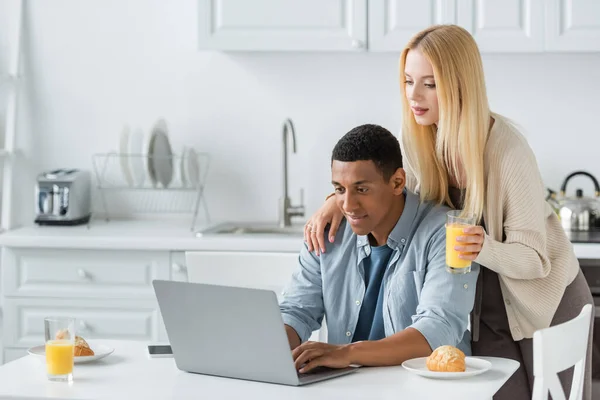 Homme afro-américain positif tapant sur ordinateur portable près du petit déjeuner et petite amie avec du jus d'orange — Photo de stock