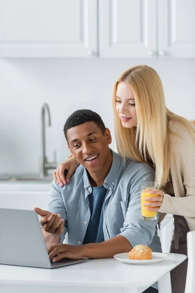 Heureux homme afro-américain pointant vers ordinateur portable près de petite amie avec du jus d'orange et croissant sur la table de cuisine — Photo de stock