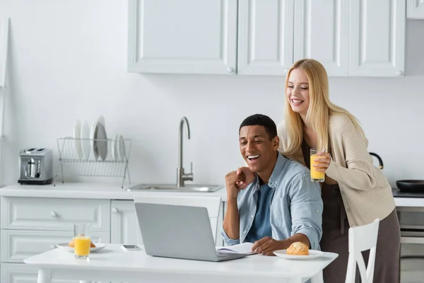 Alegre afroamericano hombre cogido de la mano con su novia mientras mira el ordenador portátil durante el desayuno - foto de stock
