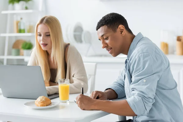 Homme afro-américain écrit dans un cahier et femme blonde travaillant sur ordinateur portable près du petit déjeuner dans la cuisine — Photo de stock