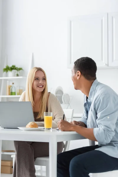 Cheerful interracial freelancers looking at each other near laptop and breakfast in kitchen — Stock Photo