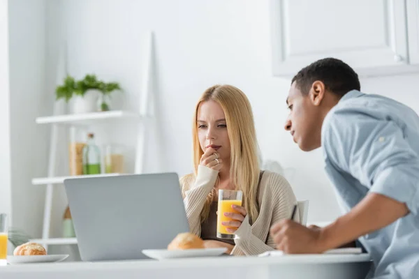 Mujer reflexiva con jugo de naranja mirando portátil cerca borrosa novio afroamericano en la cocina - foto de stock