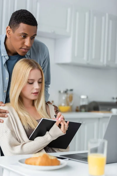 Homme afro-américain étreignant les épaules de petite amie blonde écrit dans un carnet près d'un ordinateur portable et le petit déjeuner flou — Photo de stock
