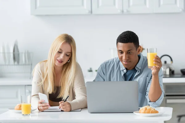 Smiling african american man with orange juice looking at laptop near girlfriend writing in notebook during breakfast — Stock Photo