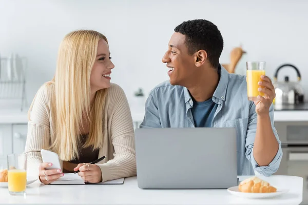 Feliz pareja interracial mirando a cada uno durante el desayuno cerca del ordenador portátil y portátil - foto de stock