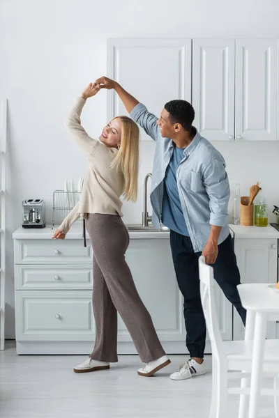 Полная длина счастливой межрасовой пары, держащейся за руки, танцуя на кухне — стоковое фото