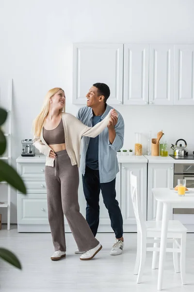 Full length of joyful interracial couple smiling at each other while dancing in modern kitchen — Stock Photo