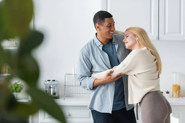 Happy african american man hugging young blonde girlfriend on blurred foreground in kitchen — Stock Photo