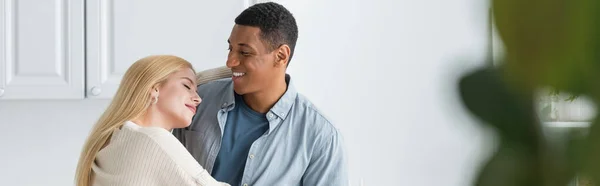 Mujer rubia feliz con los ojos cerrados abrazando sonriente novio afroamericano en la cocina, pancarta - foto de stock