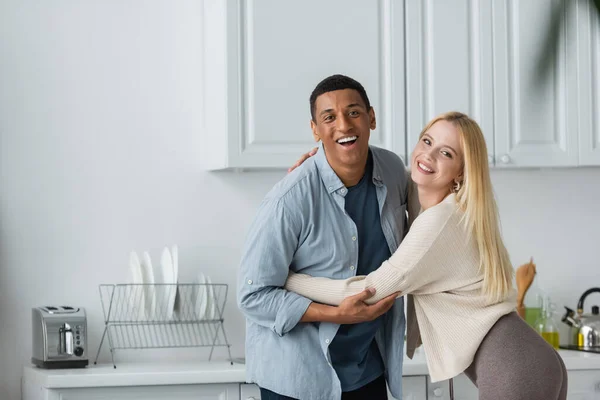 Animado interracial casal sorrindo para câmera e abraçando na cozinha — Fotografia de Stock