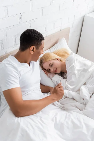 Vista de ángulo alto del hombre afroamericano mirando a la joven novia rubia durmiendo en la cama por la mañana - foto de stock