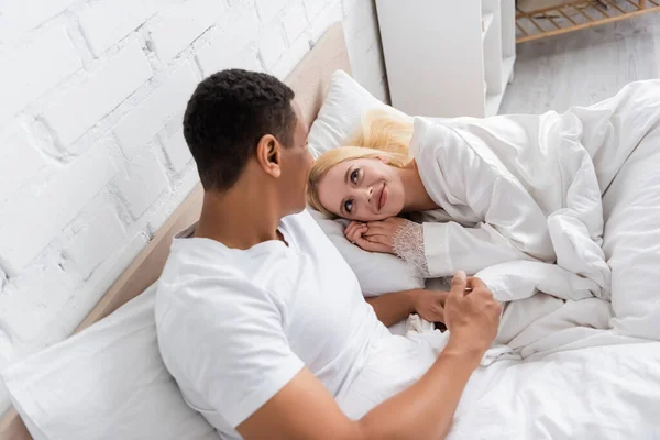 Vue grand angle de la femme blonde souriant à l'homme afro-américain tout en étant couché sur le lit le matin — Photo de stock