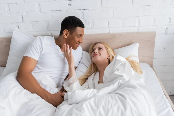 Smiling african american man looking at young and happy girlfriend touching his face on bed in morning — Stock Photo