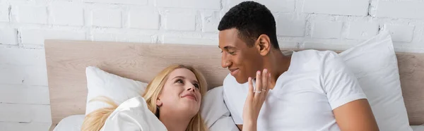 Усміхнена блондинка торкається обличчя афроамериканського хлопця, лежачи на ліжку вранці, банер — Stock Photo