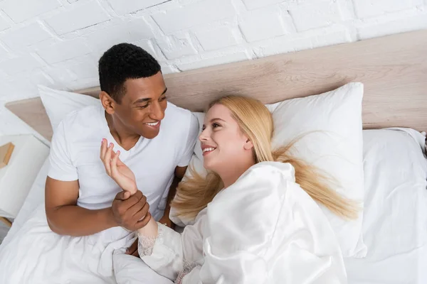 Vue grand angle de joyeux homme afro-américain tenant la main d'une jolie femme blonde sur le lit — Photo de stock