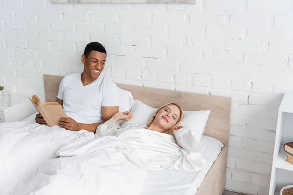 Hombre afroamericano feliz con libro mirando a novia rubia estirándose en la cama con los ojos cerrados - foto de stock