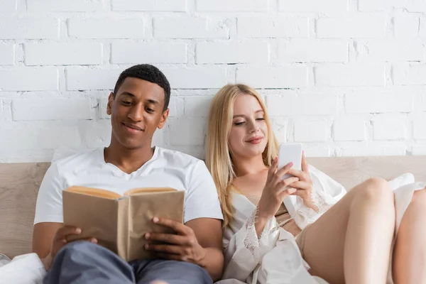 Lächelnder afrikanisch-amerikanischer Mann liest Buch neben blonder Freundin mit Smartphone im Schlafzimmer — Stockfoto