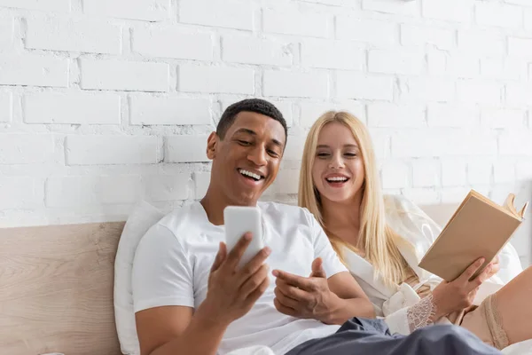 Joyful african american man pointing at cellphone near young girlfriend with book on bed — Stock Photo