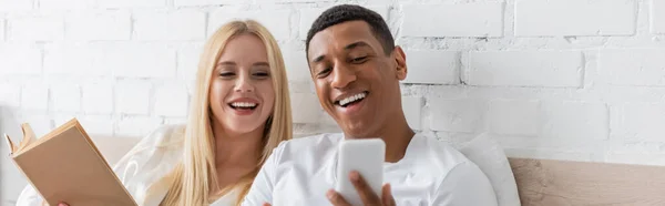 Allegro uomo afroamericano che mostra smartphone alla fidanzata sorridente che tiene libro in camera da letto, banner — Foto stock