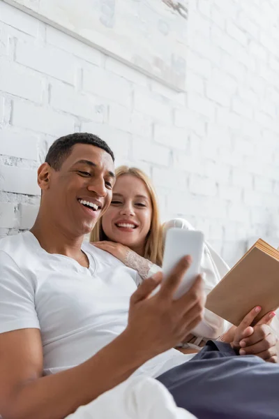 Cheerful woman with book looking at smartphone in hand of smiling african american boyfriend in bedroom — Stock Photo