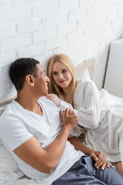 Interrazziale coppia tenendosi per mano e sorridendo a vicenda in camera da letto — Foto stock