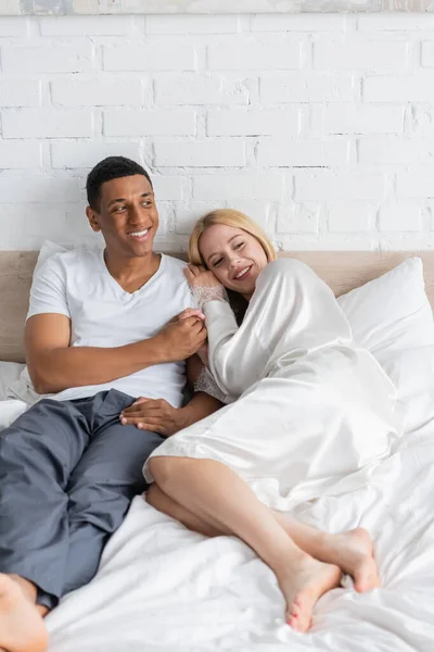 Glücklicher afrikanisch-amerikanischer Mann hält die Hand einer blonden Freundin in weißer Robe, die morgens auf dem Bett liegt — Stockfoto