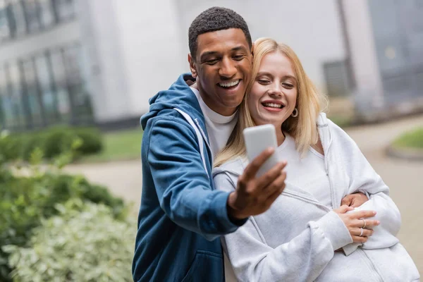 Alegre interracial pareja en sudaderas con capucha tomando selfie en móvil al aire libre - foto de stock