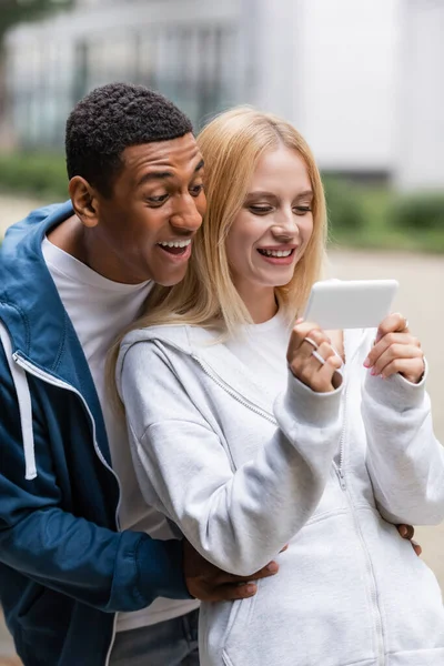 Espantado homem americano africano e mulher loira olhando para o telefone móvel ao ar livre — Fotografia de Stock