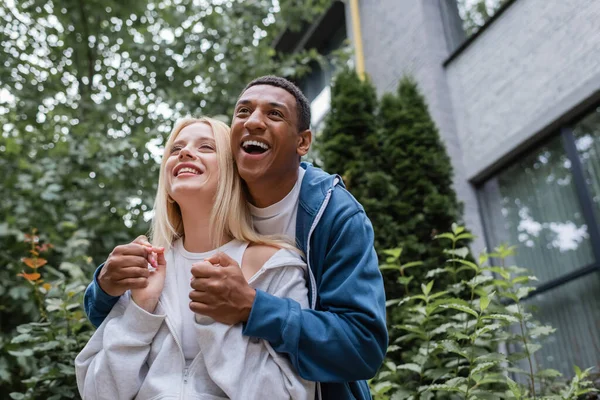 Excited african american man holding hands of blonde girlfriend and laughing on street — Stock Photo