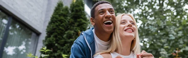 Niedrigwinkel-Ansicht aufgeregter afrikanisch-amerikanischer Mann lacht neben blonder Freundin auf der Straße der Stadt, Banner — Stockfoto