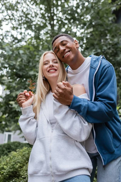 Low angle view of pleased african american man with closed eyes holding hands of young girlfriend outdoors — Stock Photo