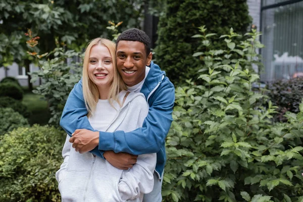 Happy african american man hugging young and cheerful girlfriend while smiling at camera — Stock Photo