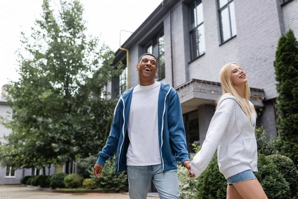 Happy interracial couple holding hands while walking near building and green plants on street — Stock Photo