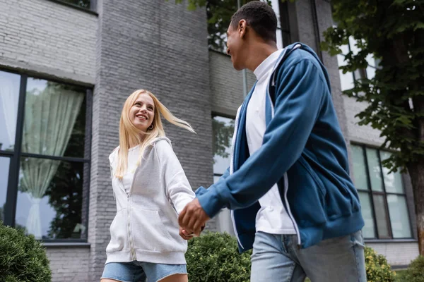 Multiethnic couple in hoodies walking on city street while holding hands and looking at each other — Stock Photo
