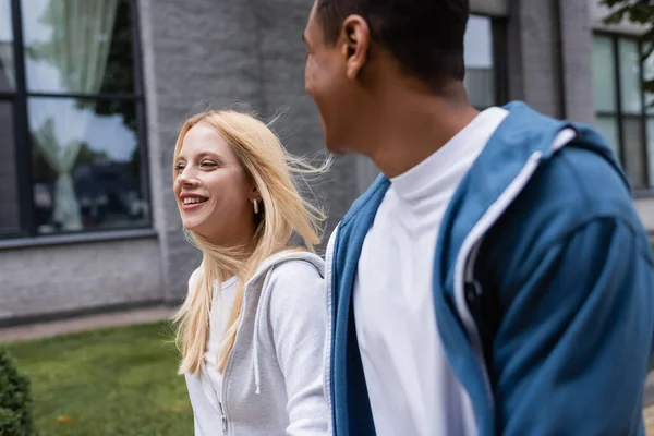 Jeune femme blonde souriant près flou afro-américain copain en plein air — Photo de stock