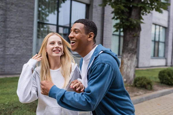Happy blonde woman looking at african american boyfriend laughing on street — Stock Photo