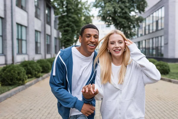 Feliz pareja multiétnica en sudaderas con capucha de la mano y mirando a la cámara en la calle urbana - foto de stock