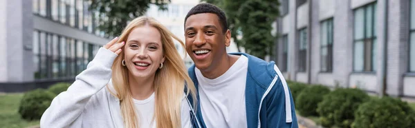 Mujer rubia y hombre afroamericano en sudaderas con capucha sonriendo a la cámara en la calle de la ciudad, bandera - foto de stock