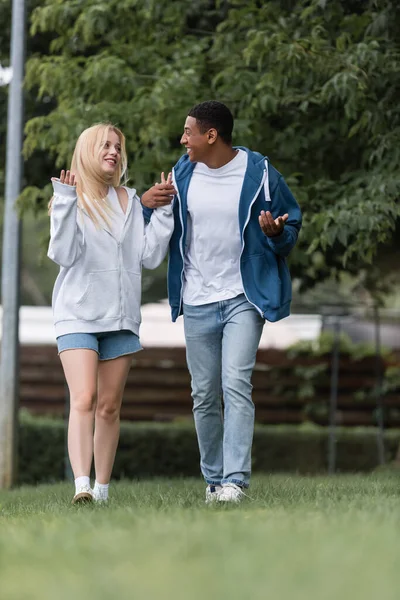 Comprimento total do casal multiétnico gesticular e falar durante a caminhada no parque — Fotografia de Stock