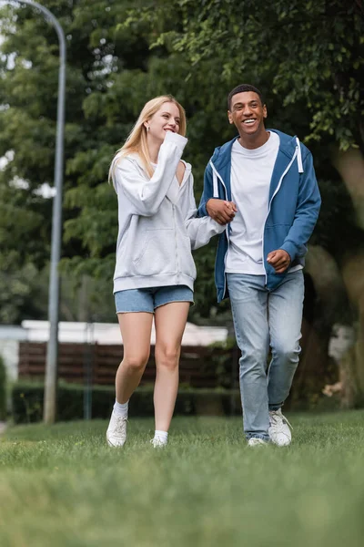 Comprimento total de casal multiétnico sorridente andando no gramado no parque e de mãos dadas — Fotografia de Stock
